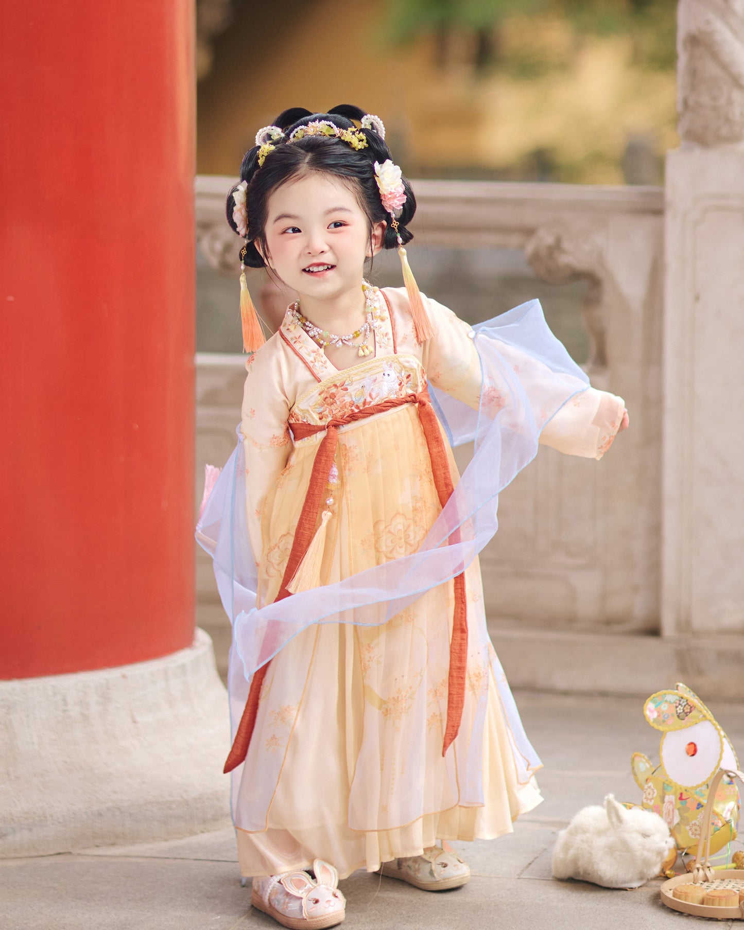Child in traditional outfit standing in front of a red column and stone wall.