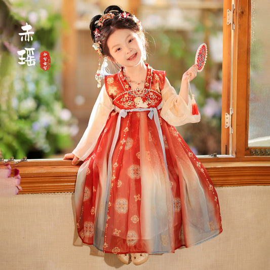 Child in a traditional red and white dress holding a toy, sitting on a windowsill with a blurred outdoor background.