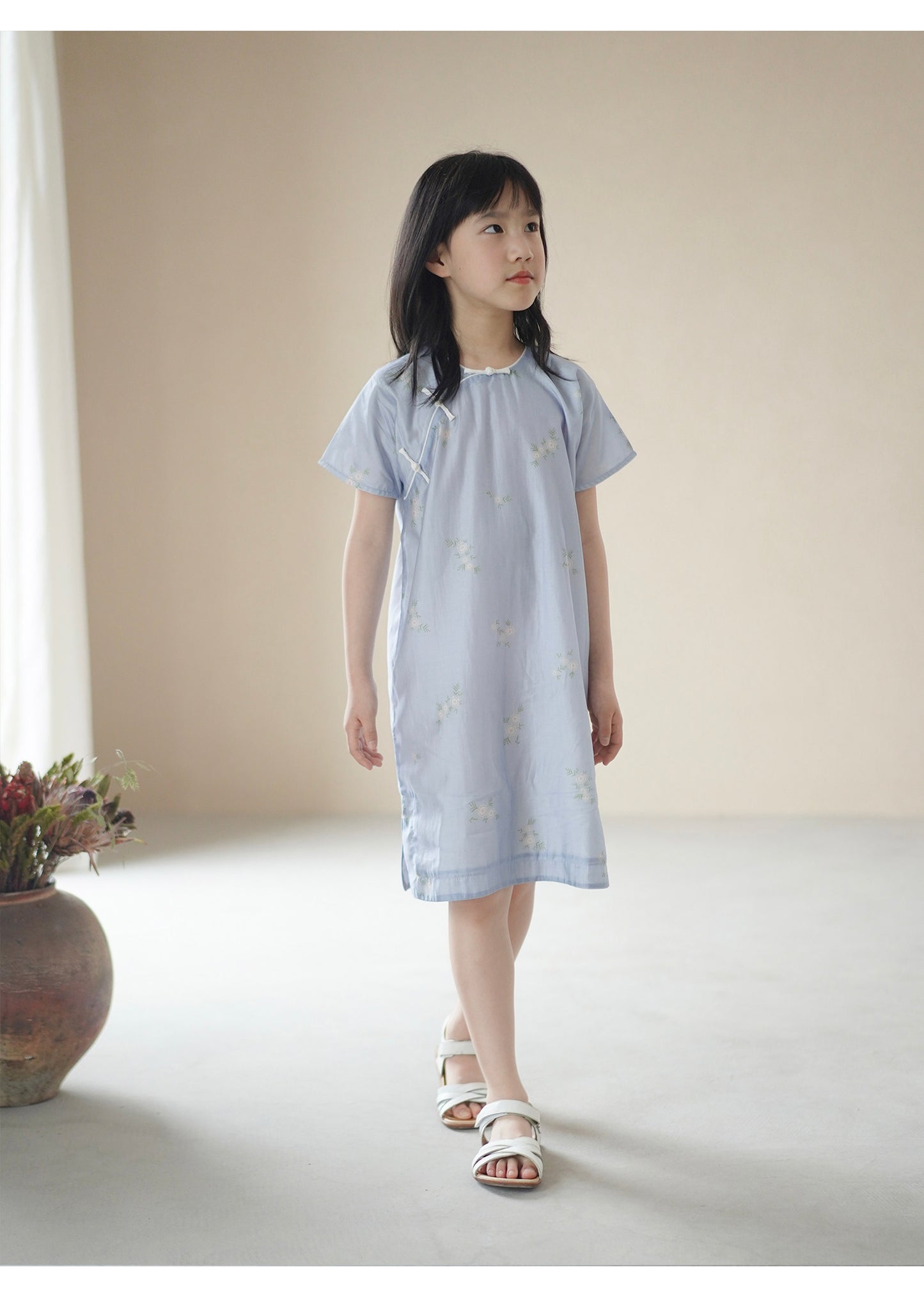 Young girl wearing a light blue dress with floral patterns in a minimalistic setting.
