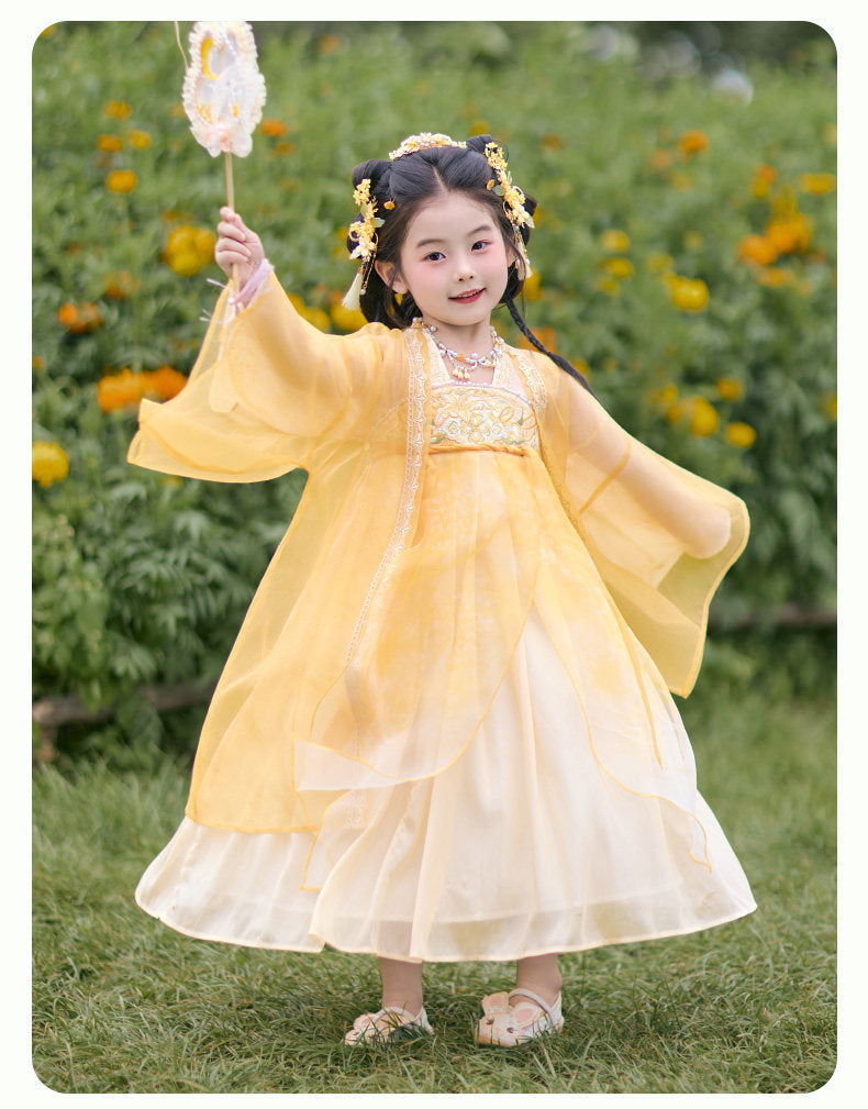Child in a yellow and white traditional outfit standing outdoors with greenery and flowers in the background.