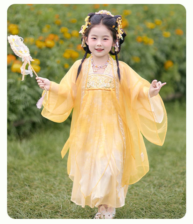 Child in a yellow traditional outfit with floral headpiece and fan, standing outdoors.