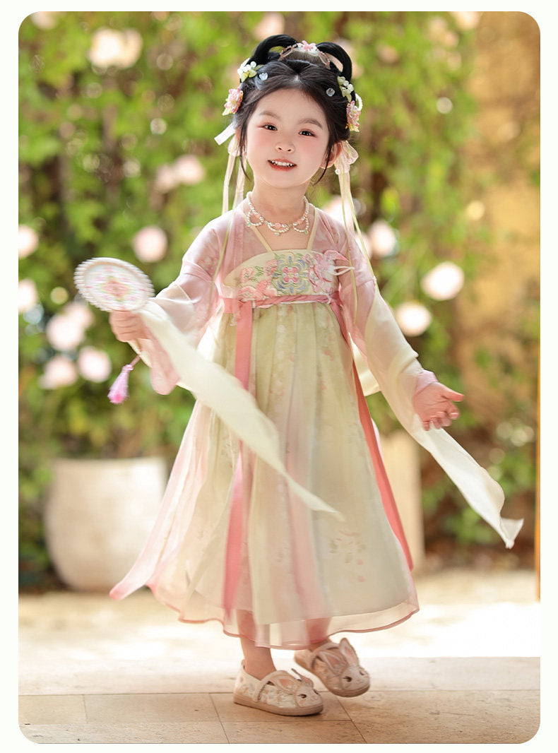 Child in traditional outfit with floral hair accessories and a white ribbon outdoors.