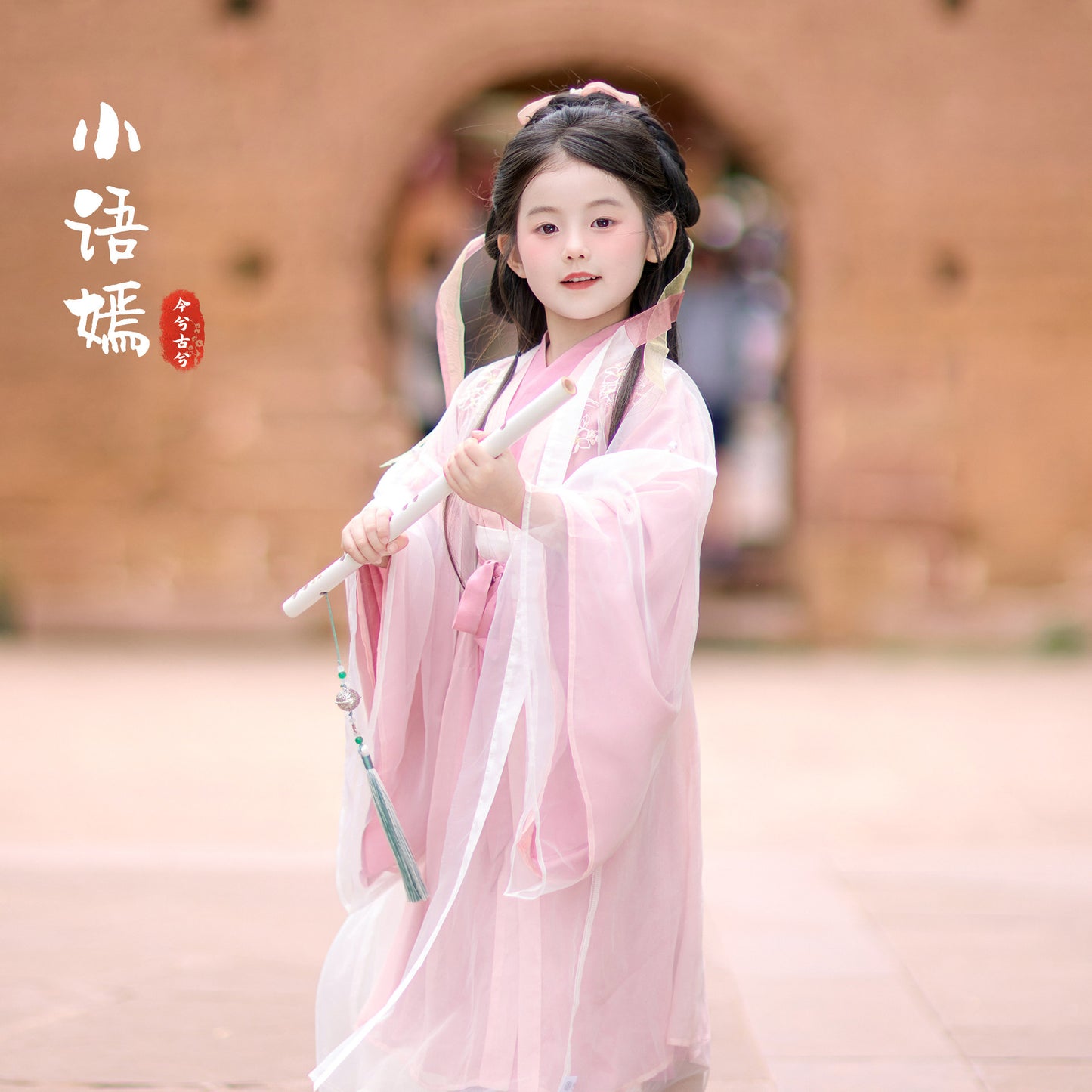 Young girl in a pink traditional outfit holding a staff in front of a blurred architectural background