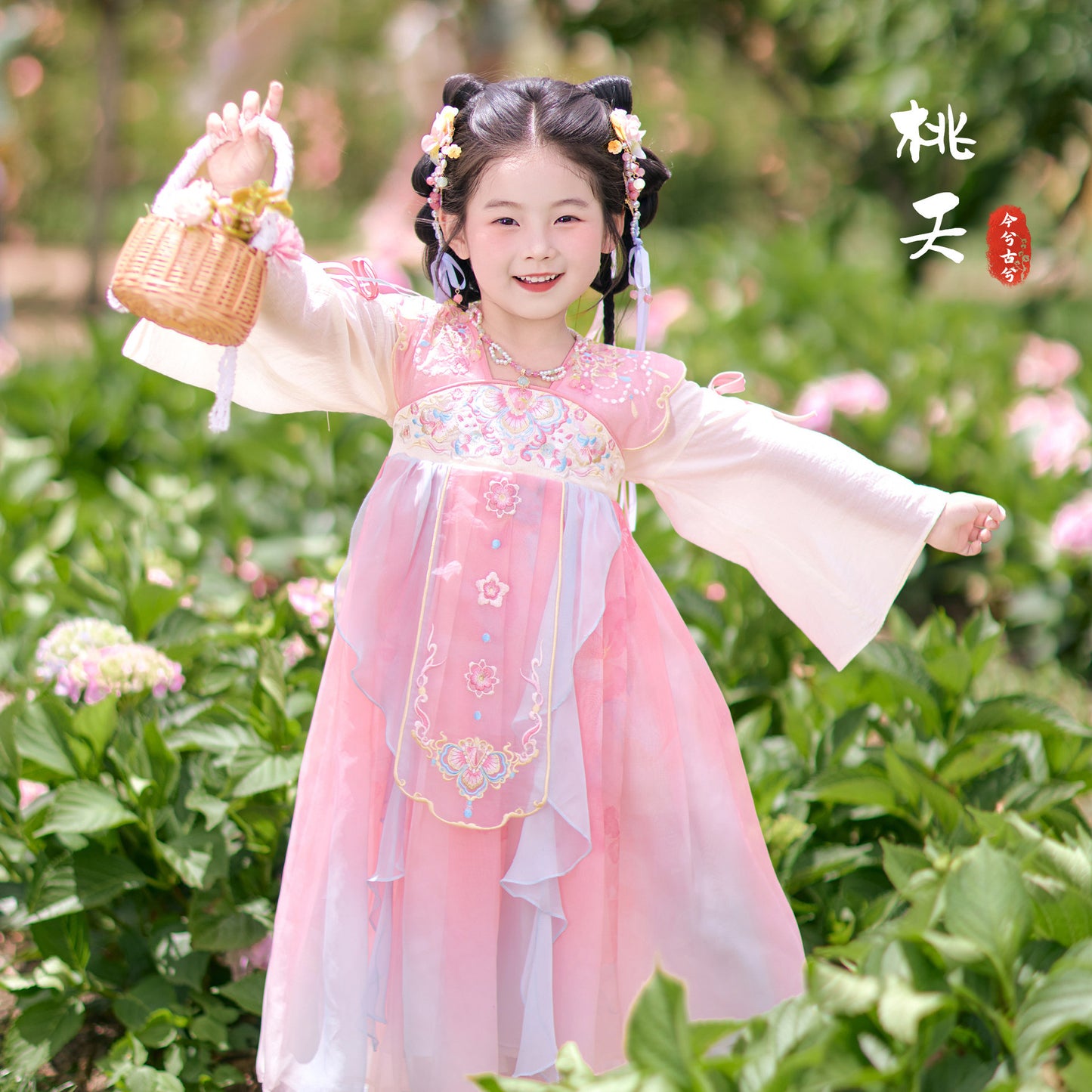 Child in traditional pink and white dress standing in a garden with flowers and greenery.