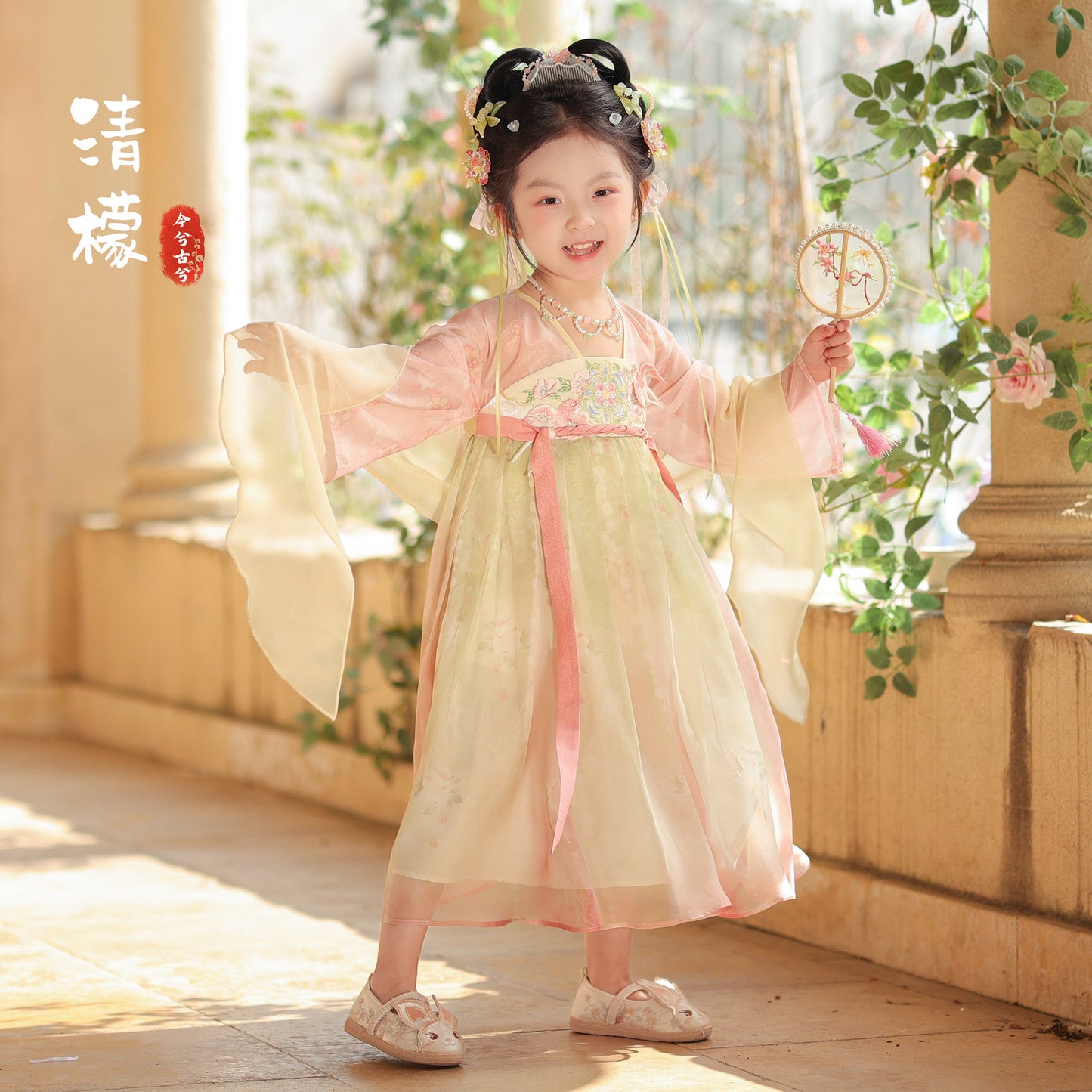 Child in traditional outfit with floral hair accessories and a fan, standing in a sunlit room with columns and plants.
