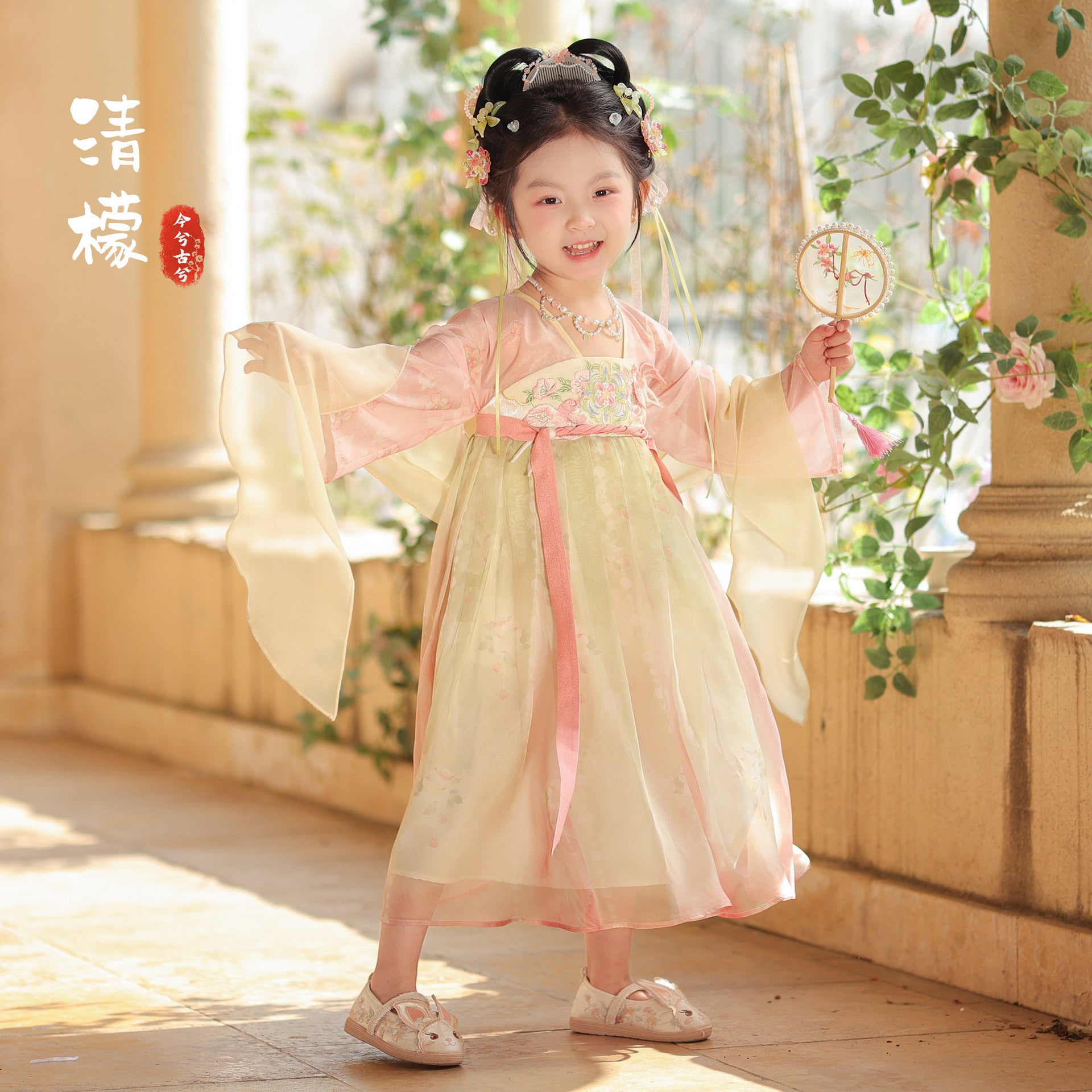 Child in traditional outfit with floral hair accessories and a fan, standing in a sunlit room with columns and plants.