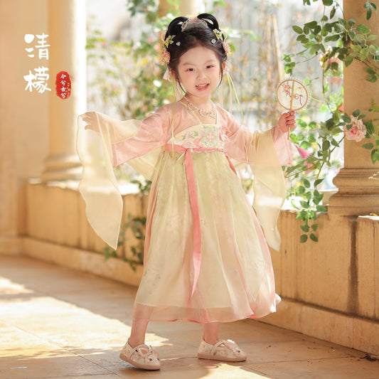 Child in traditional outfit with floral hair accessories and a fan, standing in a sunlit room with columns and plants.