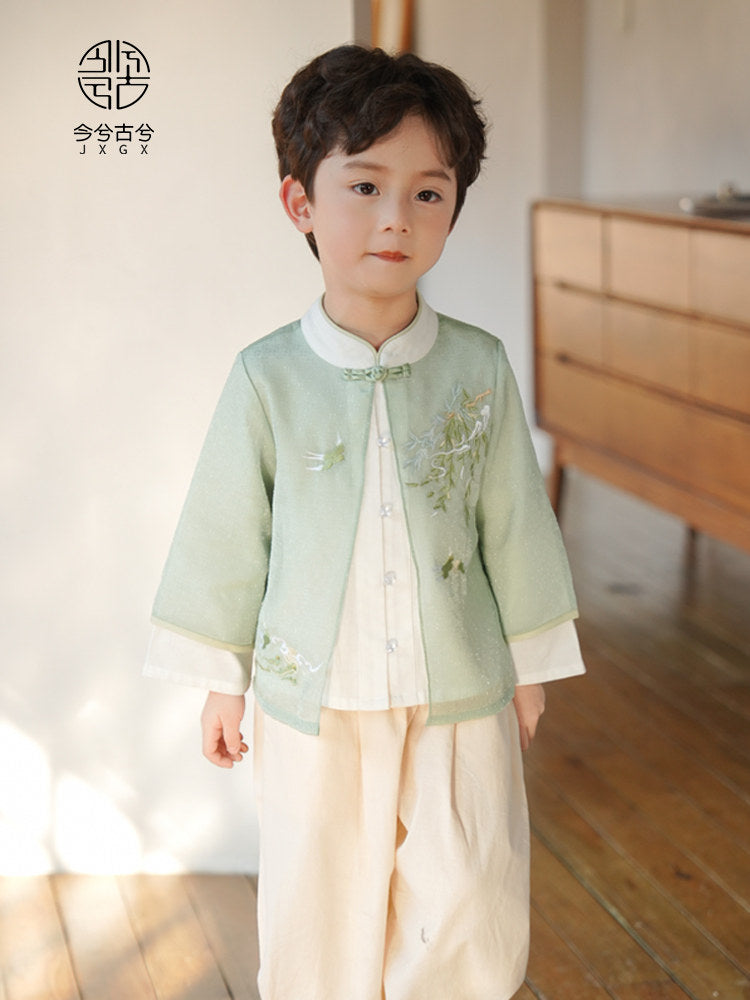 Child wearing a light green traditional outfit with floral embroidery in a room with wooden floor and white wall.
