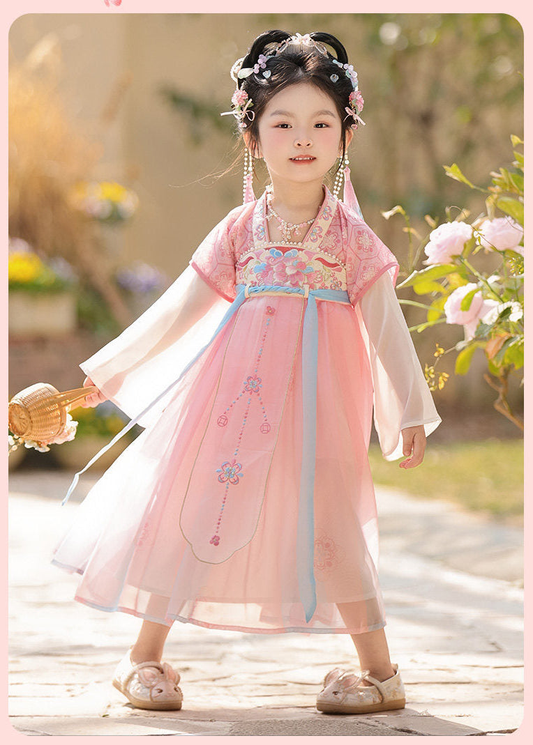 Child in traditional pink dress with floral headpiece standing outdoors.