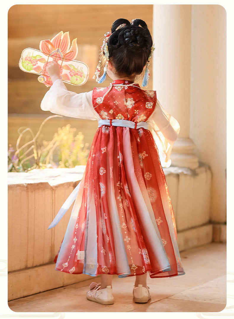 Child in a traditional red and white dress holding a decorative fan indoors.