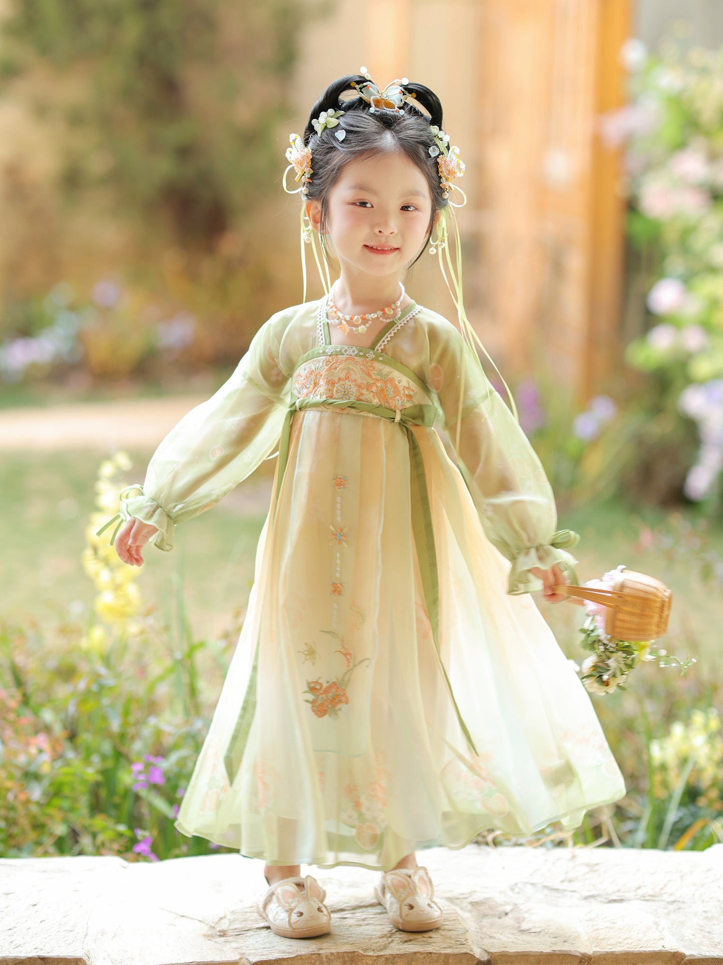 Young girl in a traditional light green dress standing outdoors with flowers and greenery in the background.