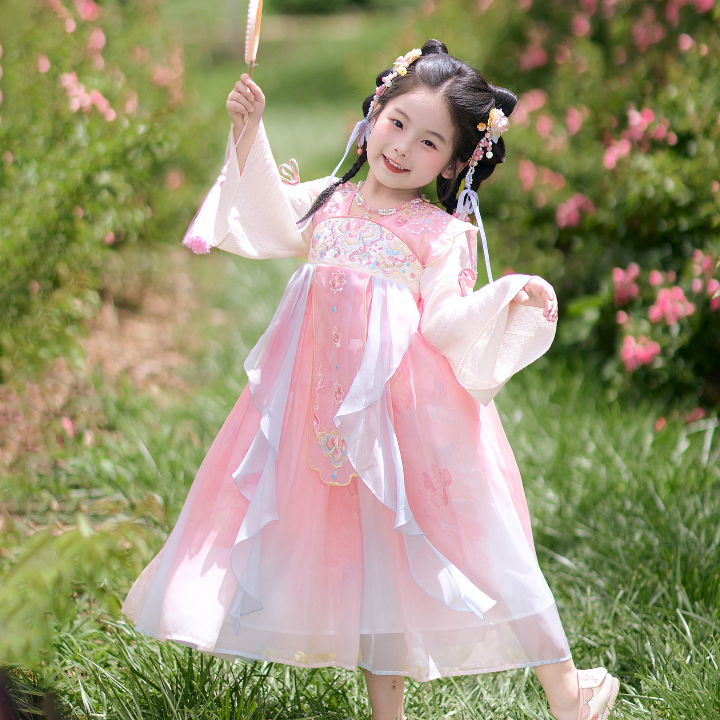 Child in a pink and white traditional outfit standing in a garden with green grass and pink flowers.