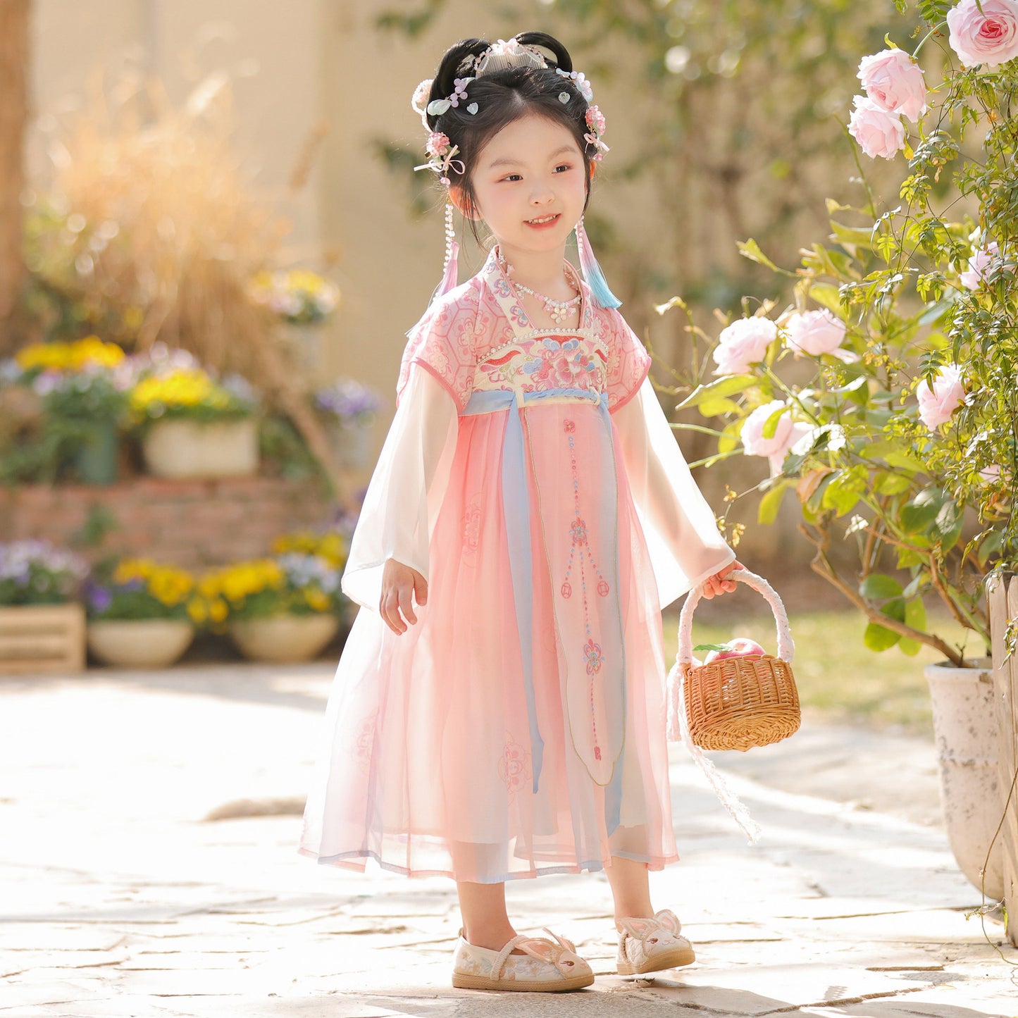 Young girl in a traditional pink dress holding a basket, standing in a garden with flowers and plants.