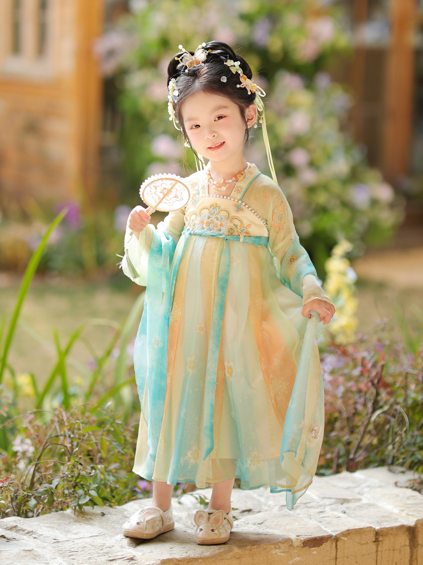 Child in traditional outfit standing outdoors with flowers and wooden structure in the background
