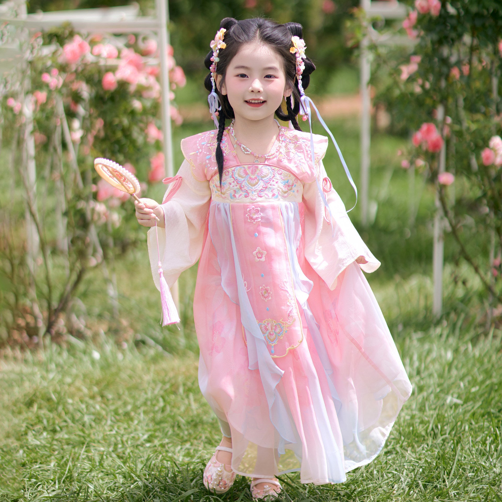 Young girl in a pink traditional dress standing in a garden with flowers and greenery.