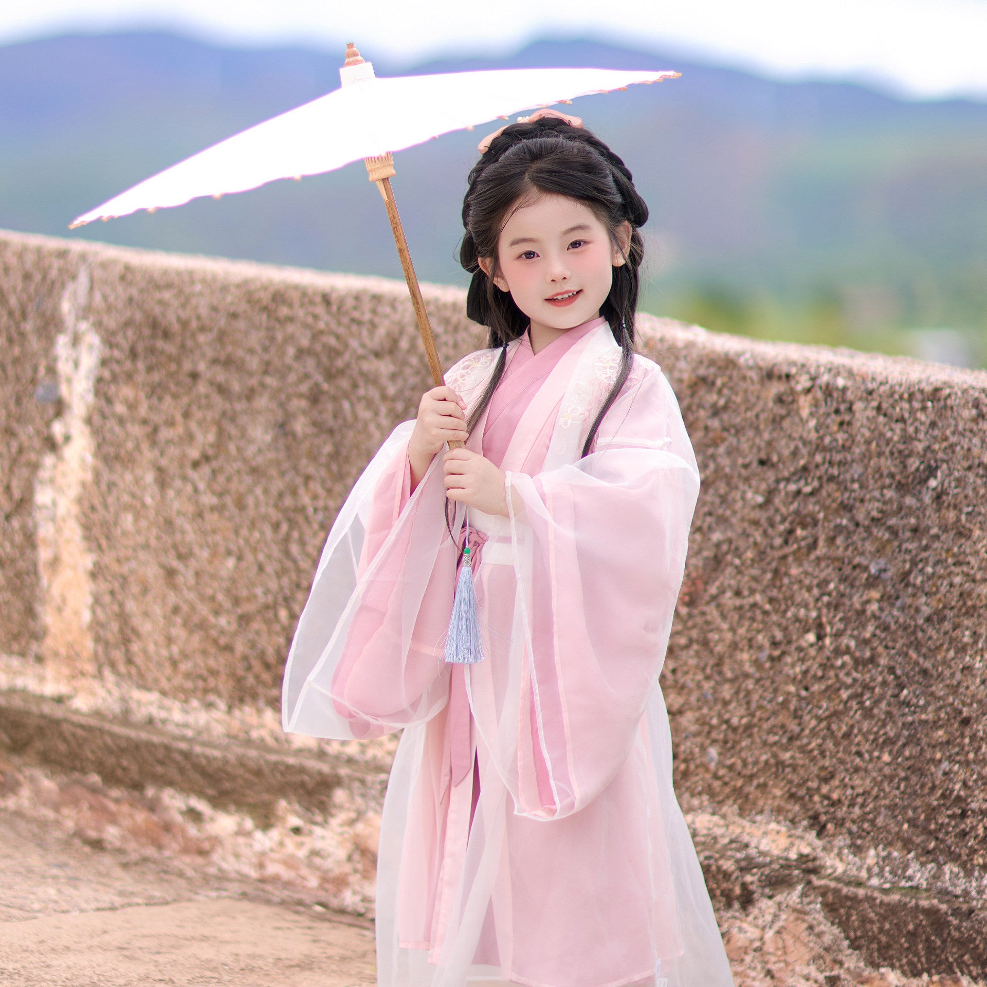 Child in traditional pink and white outfit holding an umbrella with a scenic background