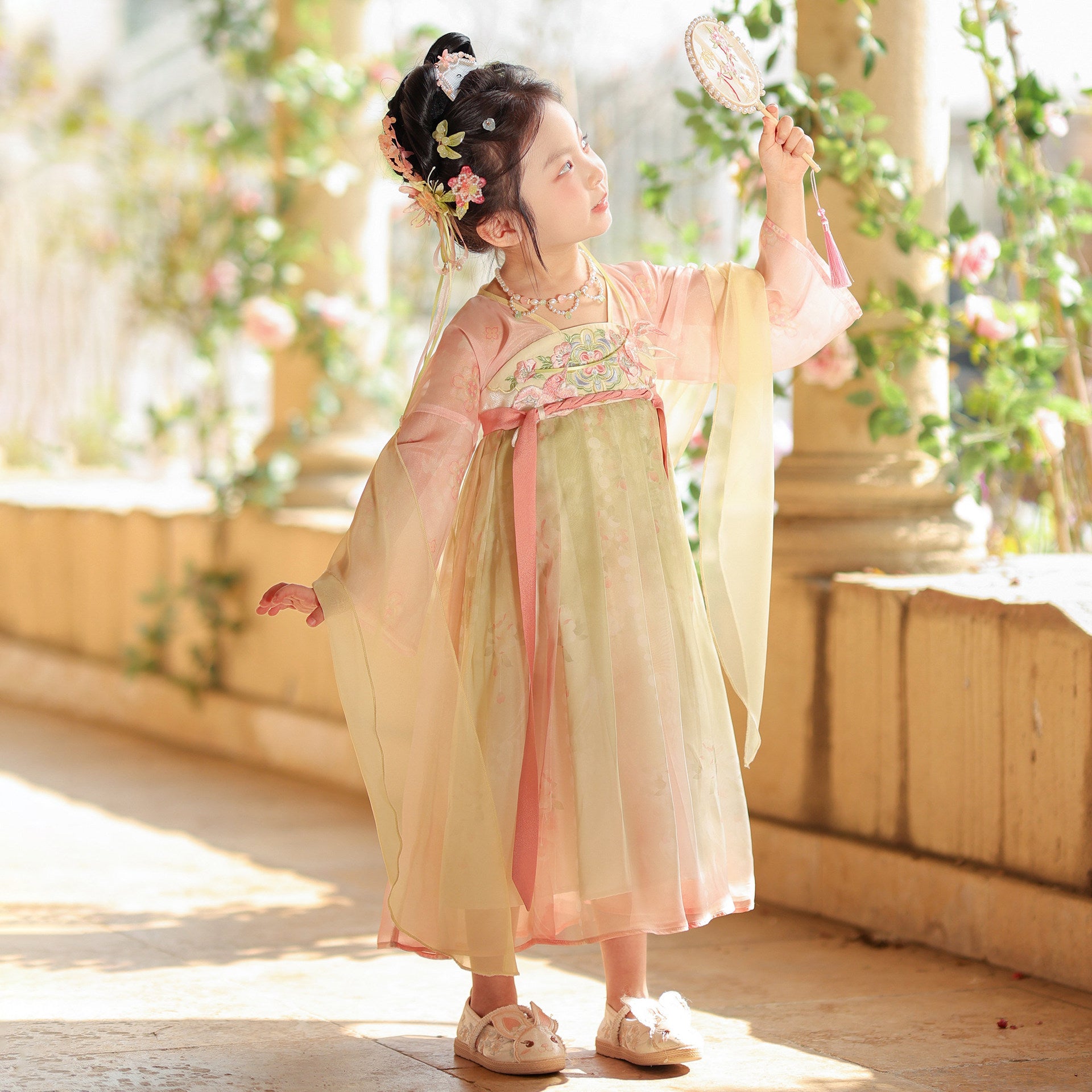 Young girl in a traditional outfit holding a fan, standing in a softly lit room with plants.