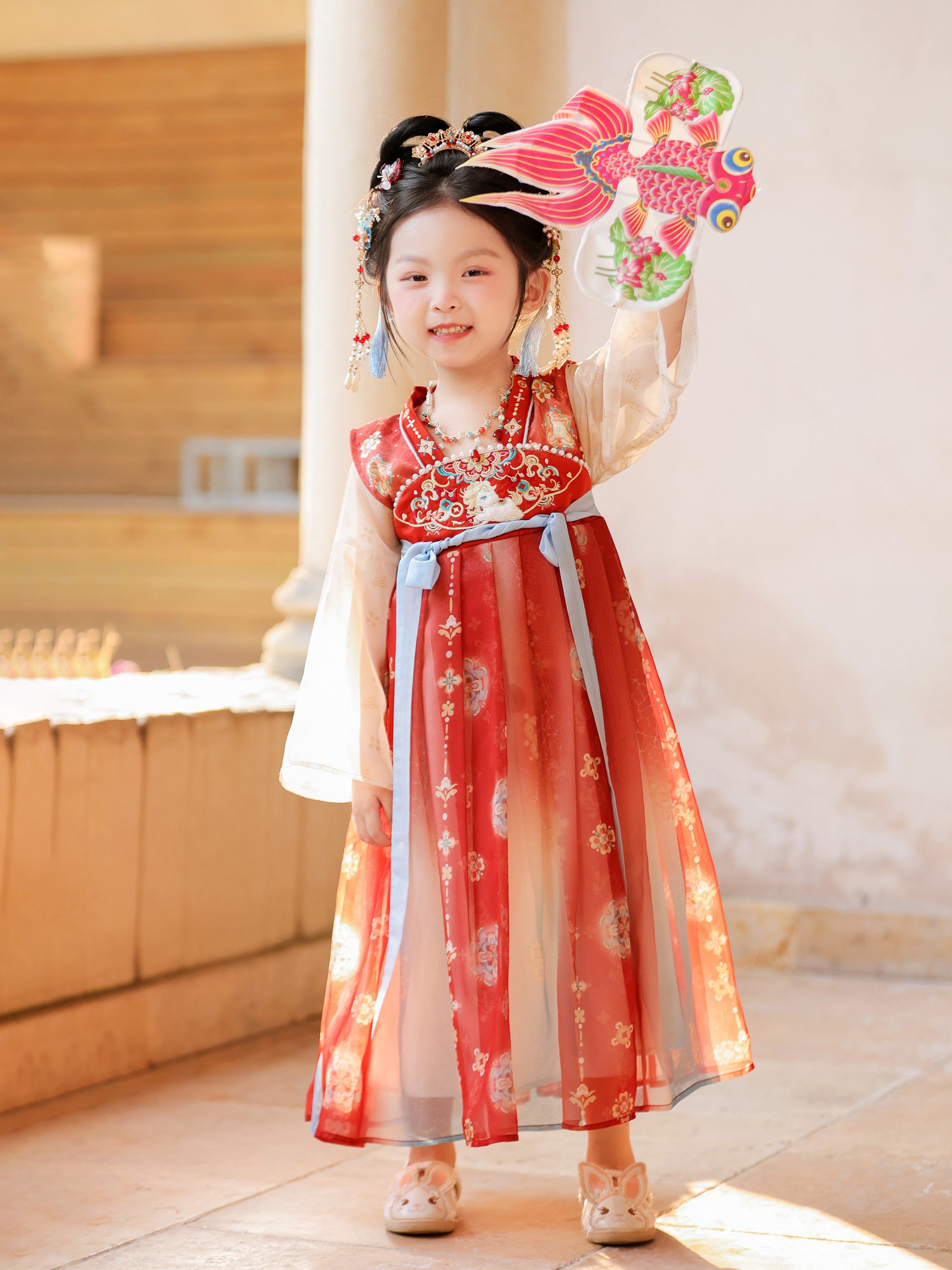 Child in a traditional red dress holding a colorful fan indoors.