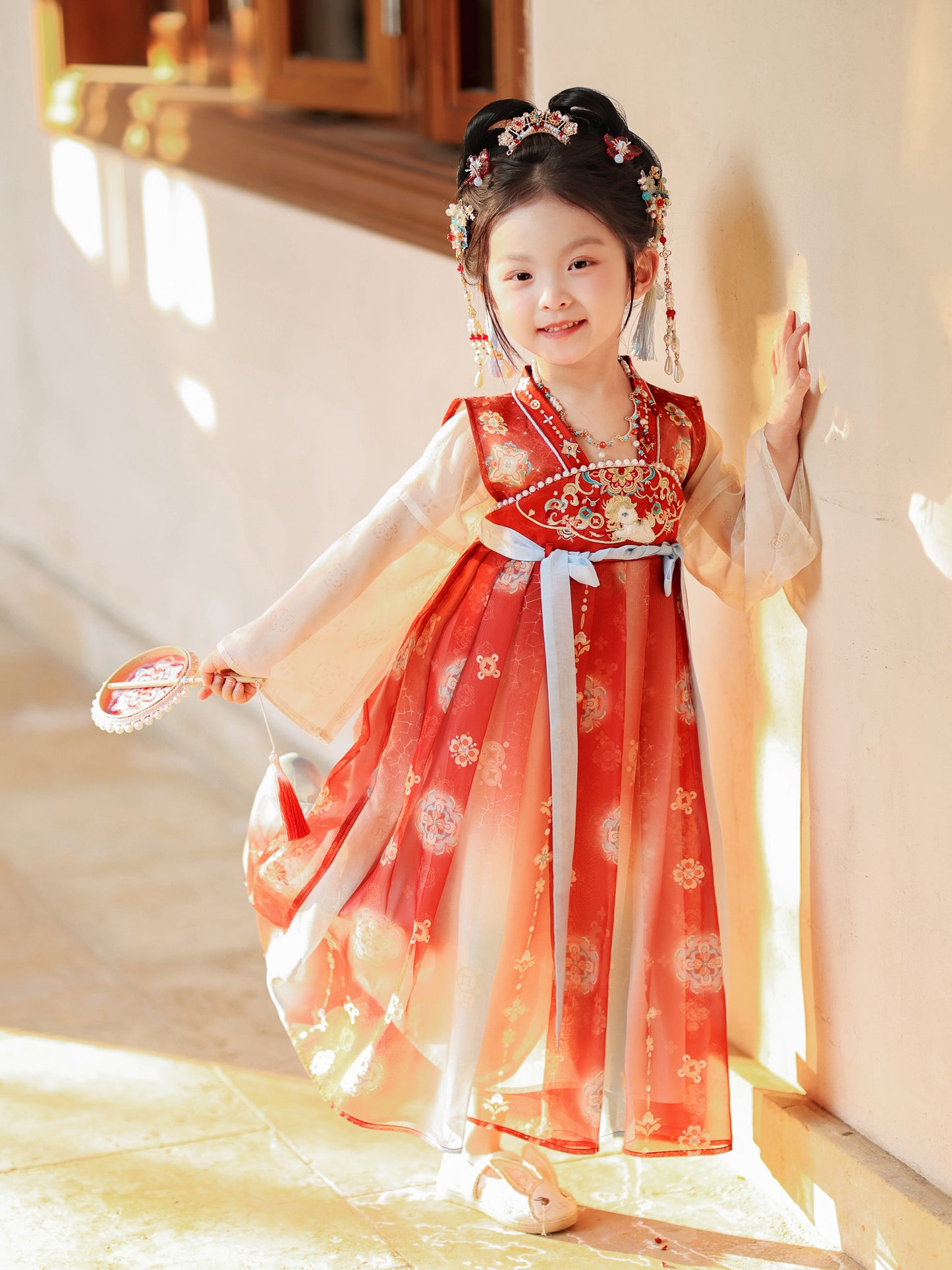 Child in a traditional red and white dress with floral hair accessories, standing indoors.