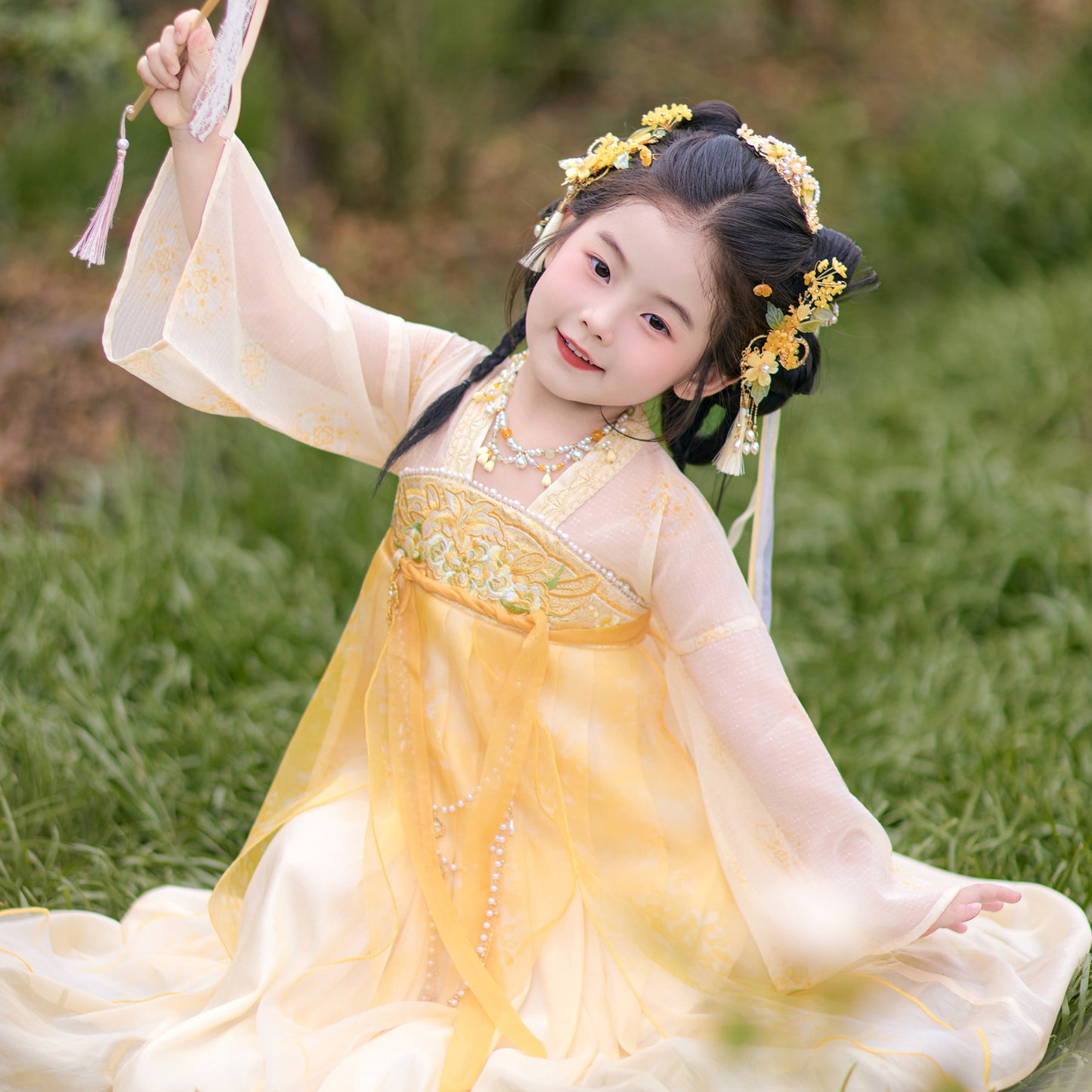 Young girl in a traditional yellow and white dress with floral hair accessories sitting on grass.