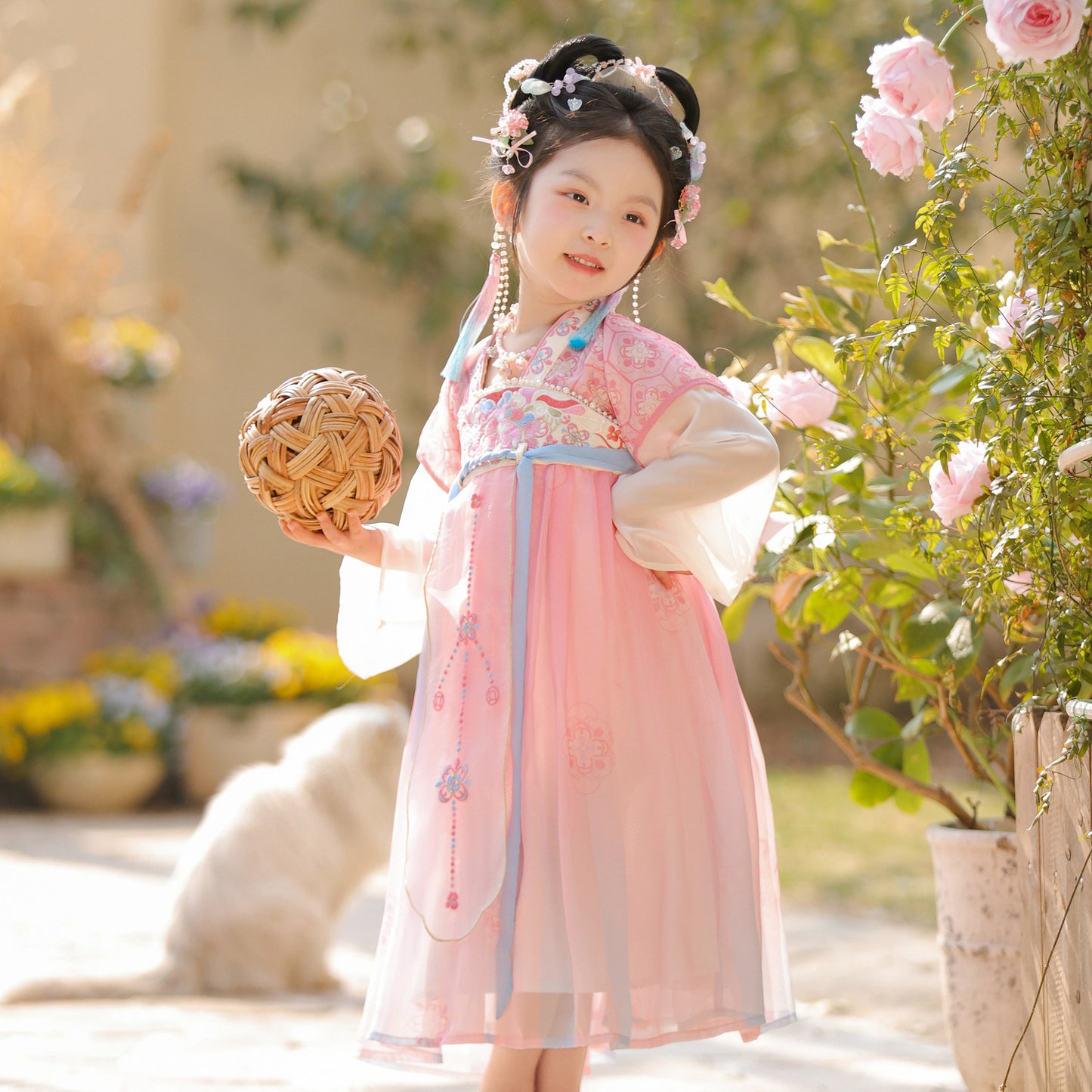 Young girl in traditional pink dress holding a woven ball, standing in a garden with flowers and plants.
