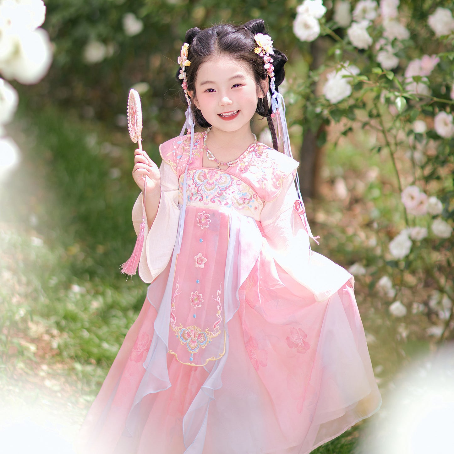 Young girl in a traditional pink dress holding a fan, standing in a garden with white flowers.