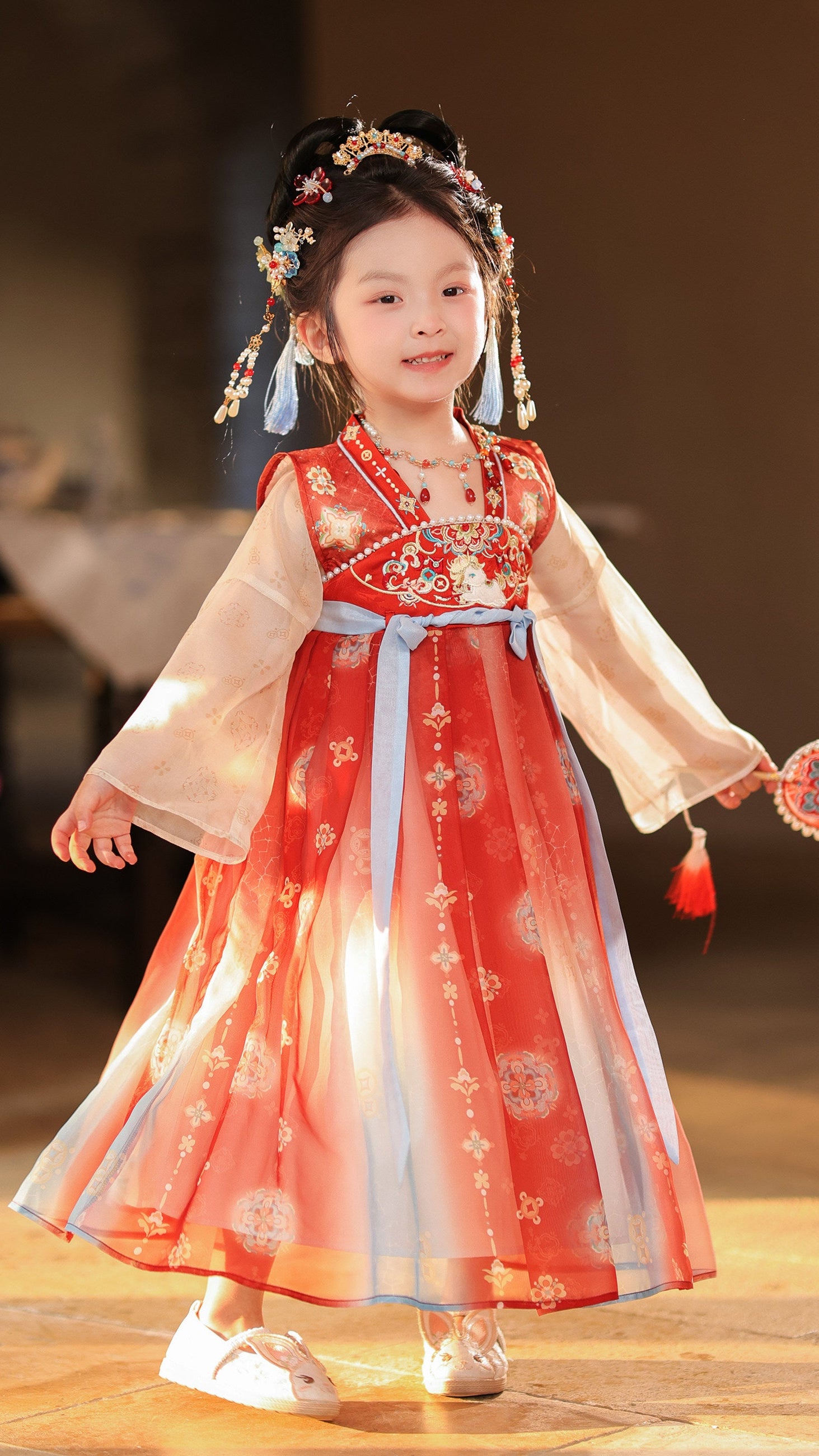 Child in traditional red and white dress with floral hair accessories on a wooden floor.
