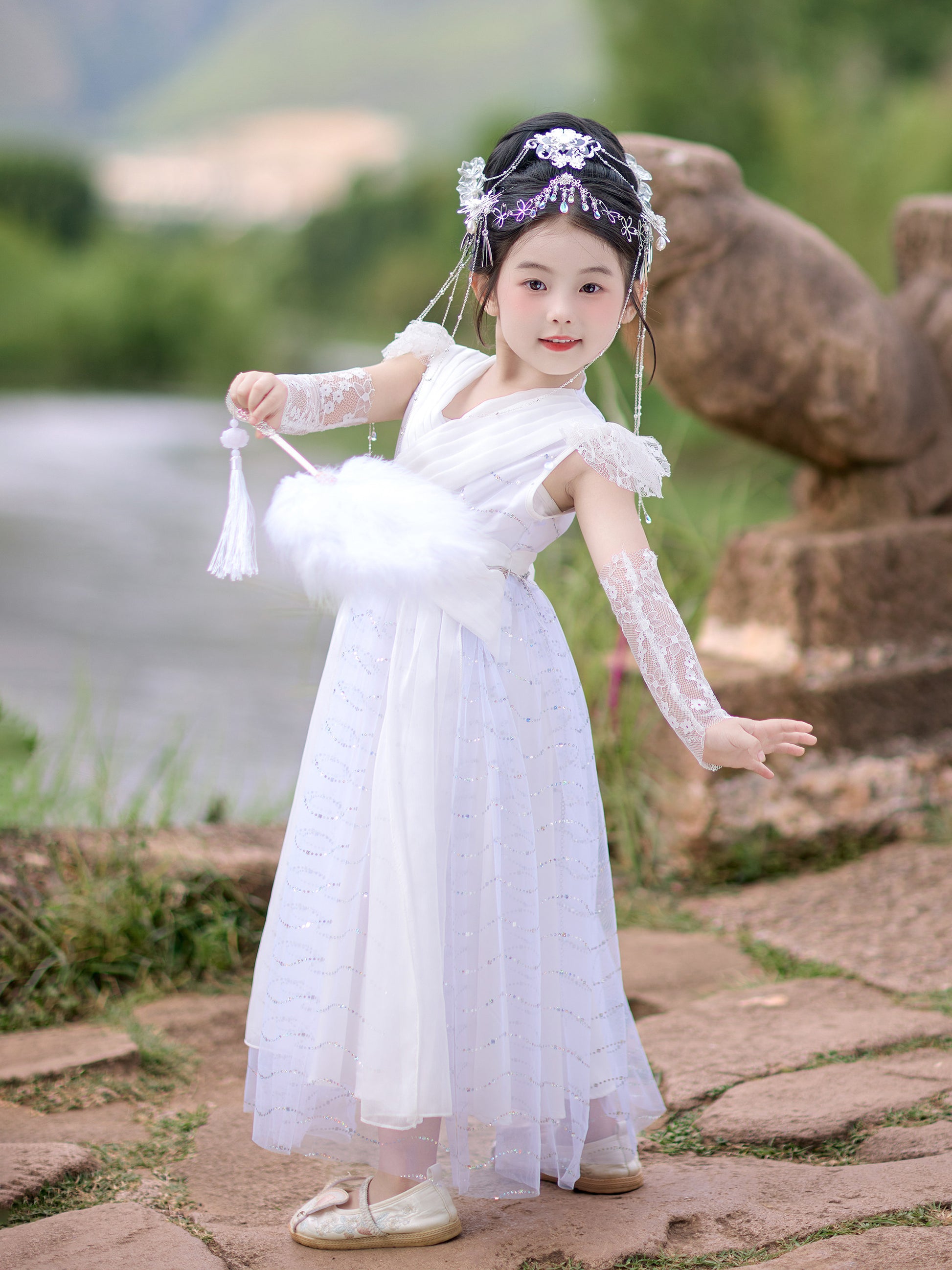 Young girl in a white dress with decorative headpiece standing outdoors near a body of water.