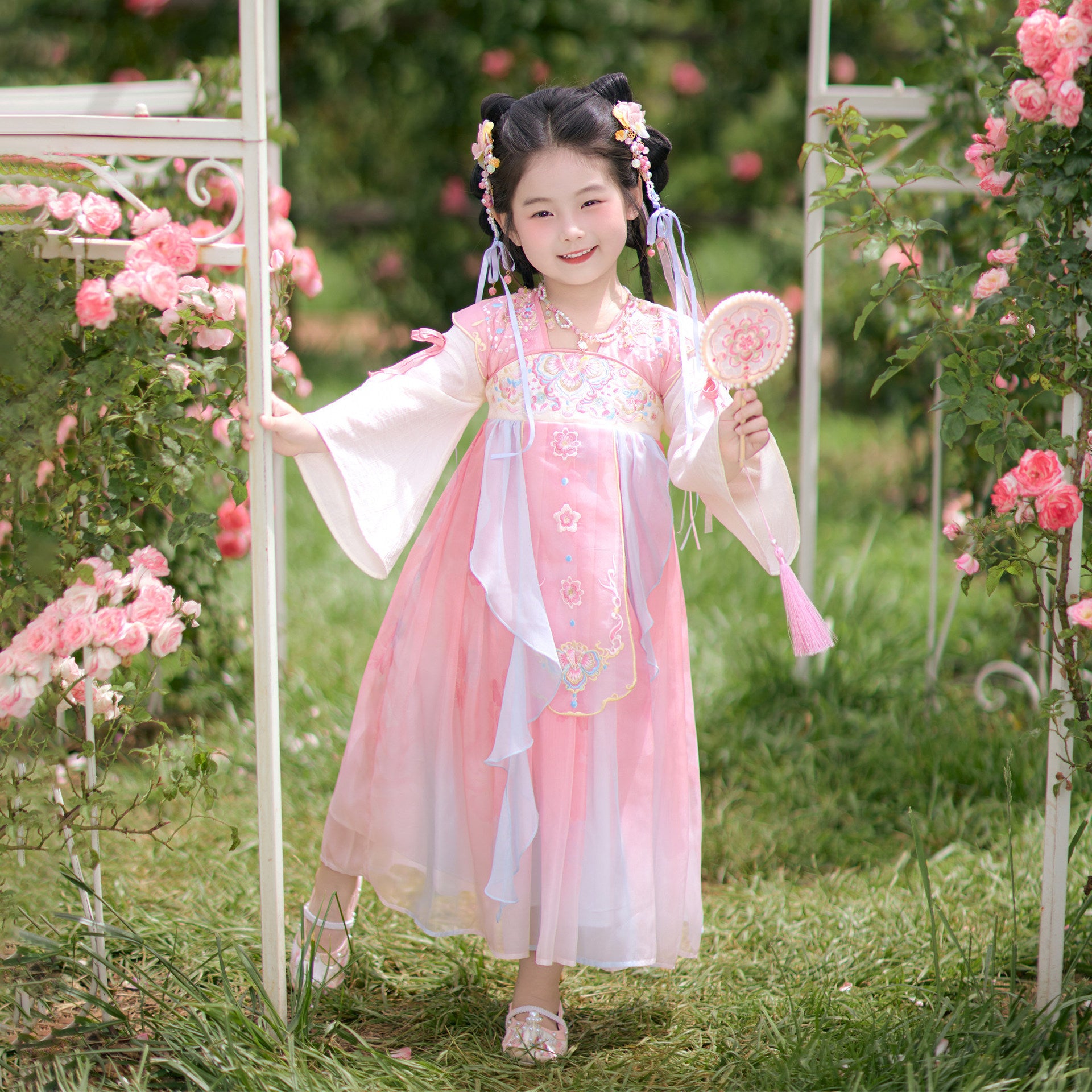 Young girl in a traditional pink and white dress standing in a garden with pink flowers.