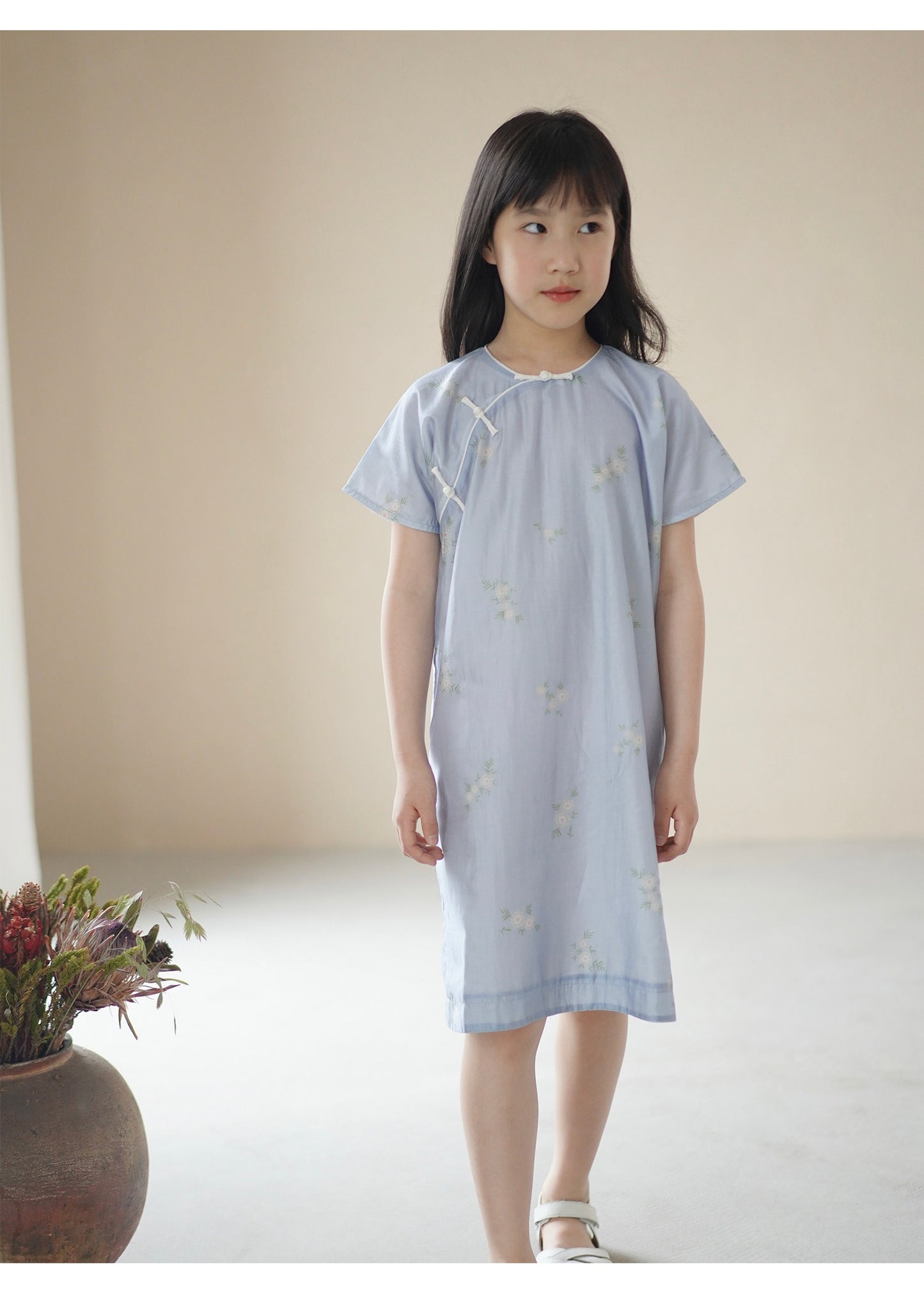 Young girl wearing a light blue dress with a subtle pattern, standing against a plain background.