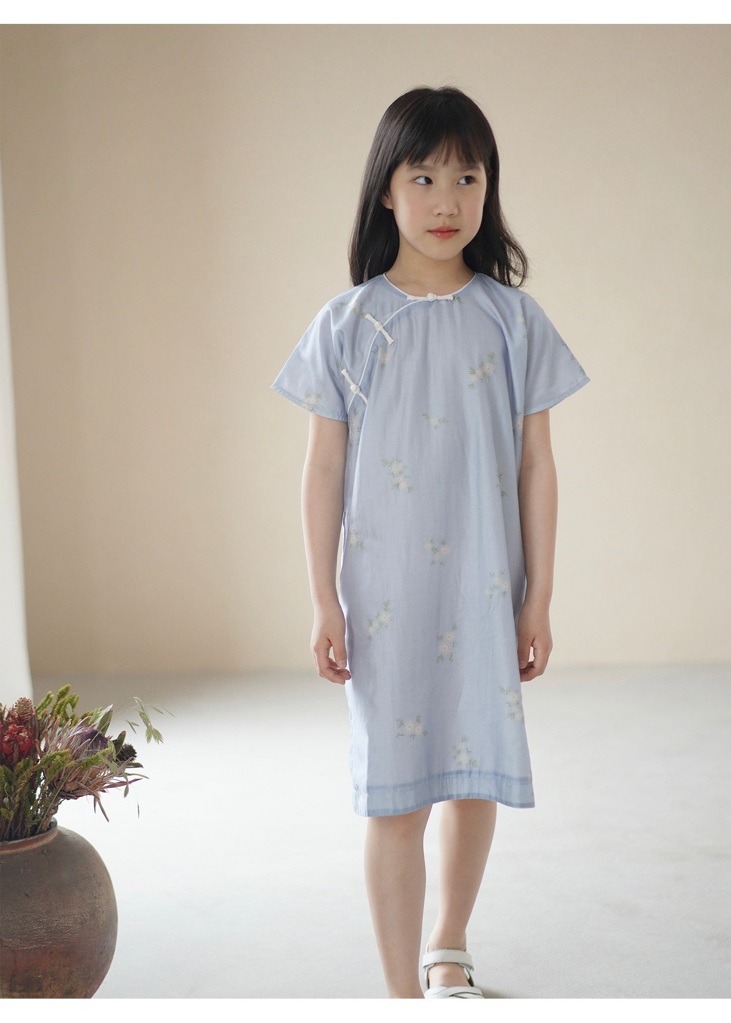 Young girl wearing a light blue dress with a subtle pattern, standing against a plain background.