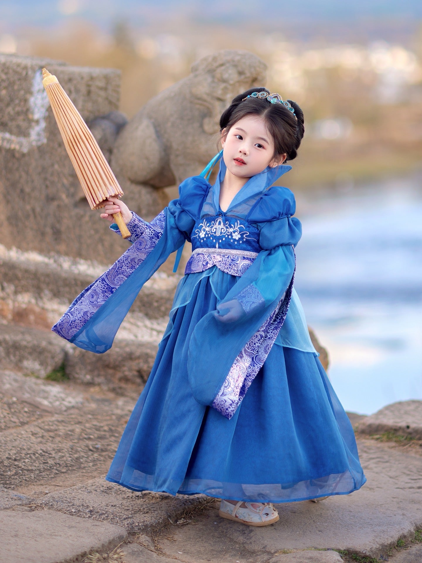 Child in a blue traditional outfit holding a bamboo umbrella with a blurred natural background