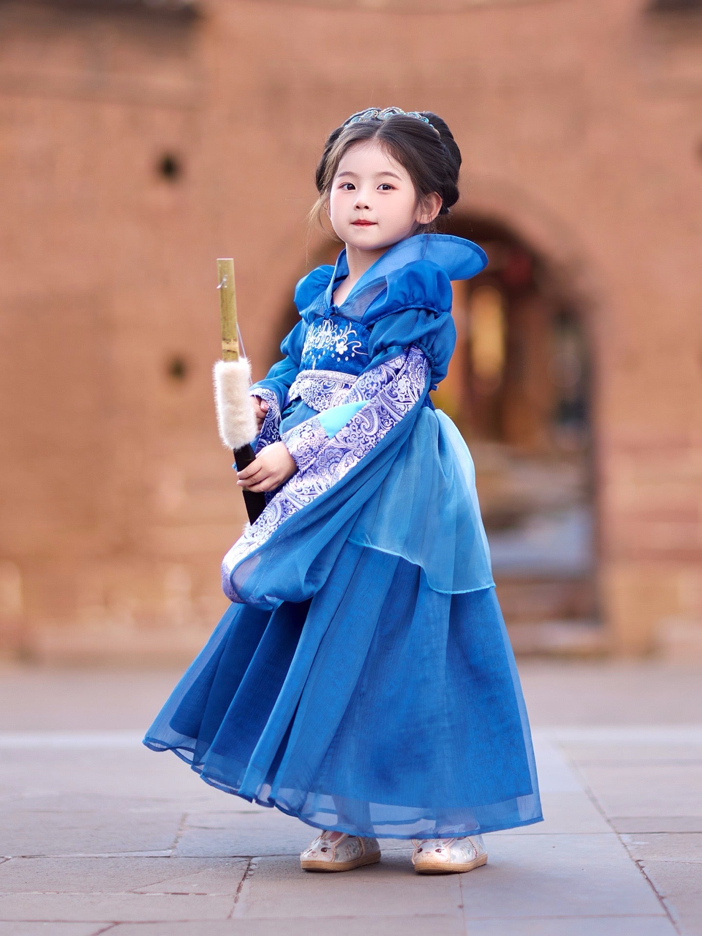 Child in a blue traditional outfit standing in front of a brick wall.
