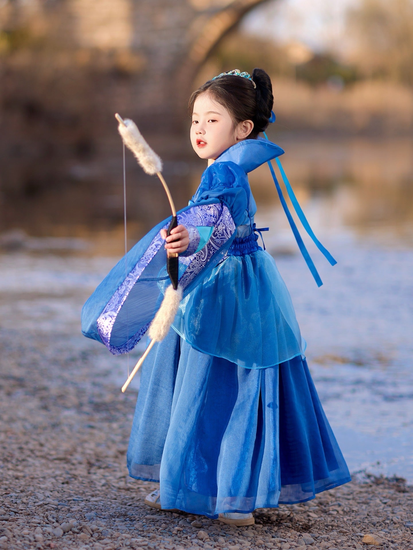 Child in a blue traditional outfit holding a decorative staff outdoors.