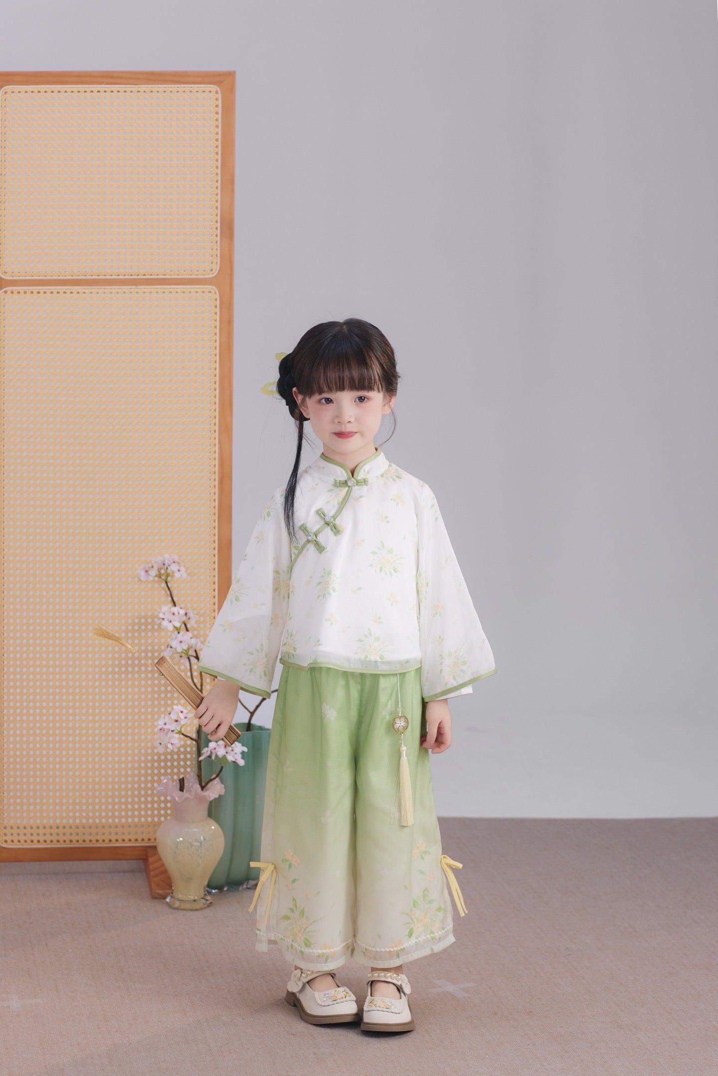 Child in traditional outfit standing in a room with a light gray wall and wooden screen.