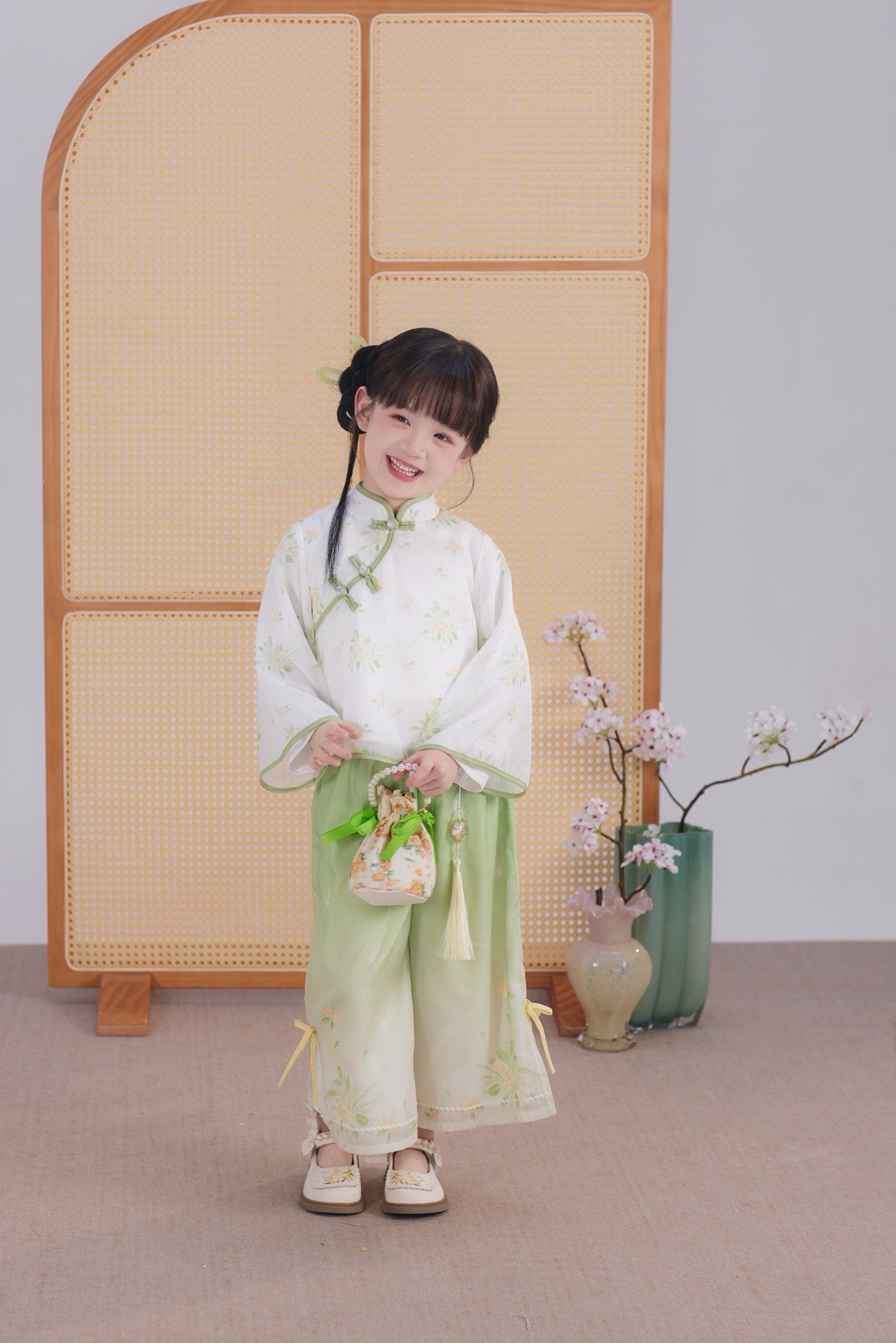 Child in traditional outfit standing in a room with a woven screen and decorative vases.