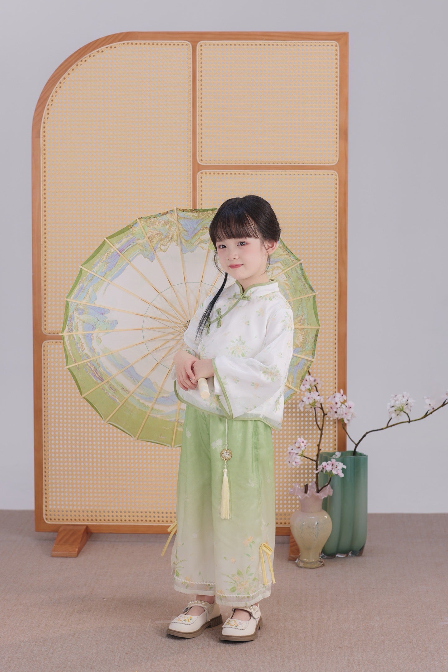 Child in traditional green and white outfit holding a decorative fan against a light background