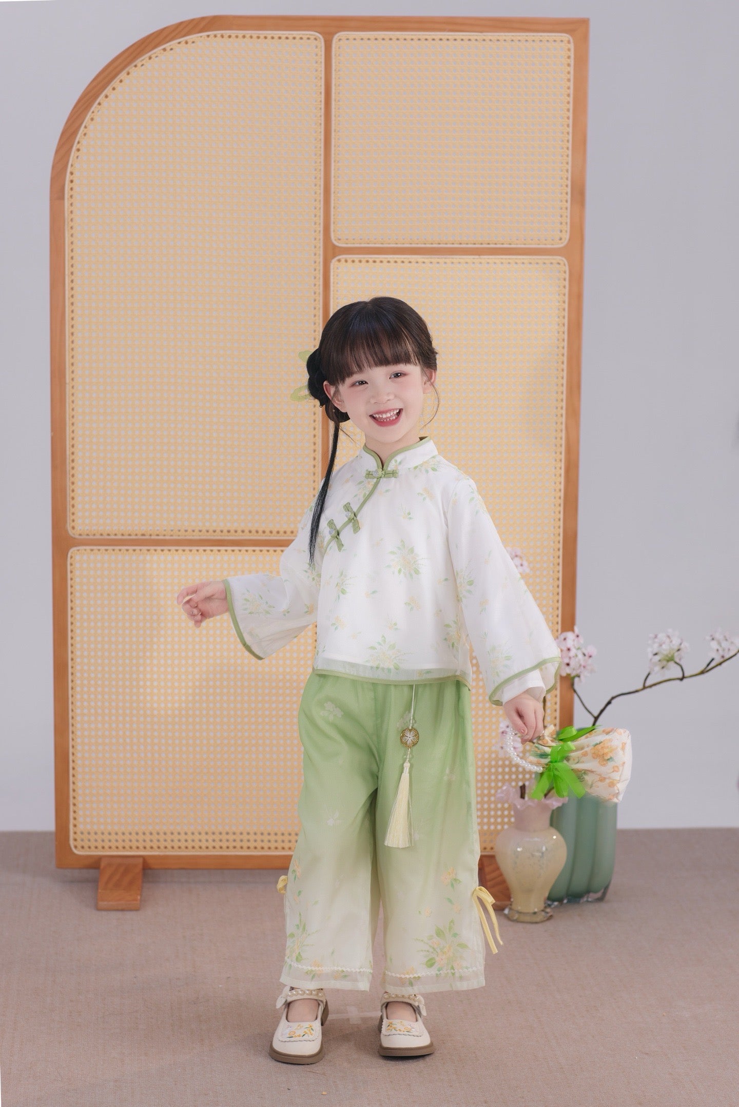 Child in traditional outfit standing in front of a wooden screen with a vase and flowers.