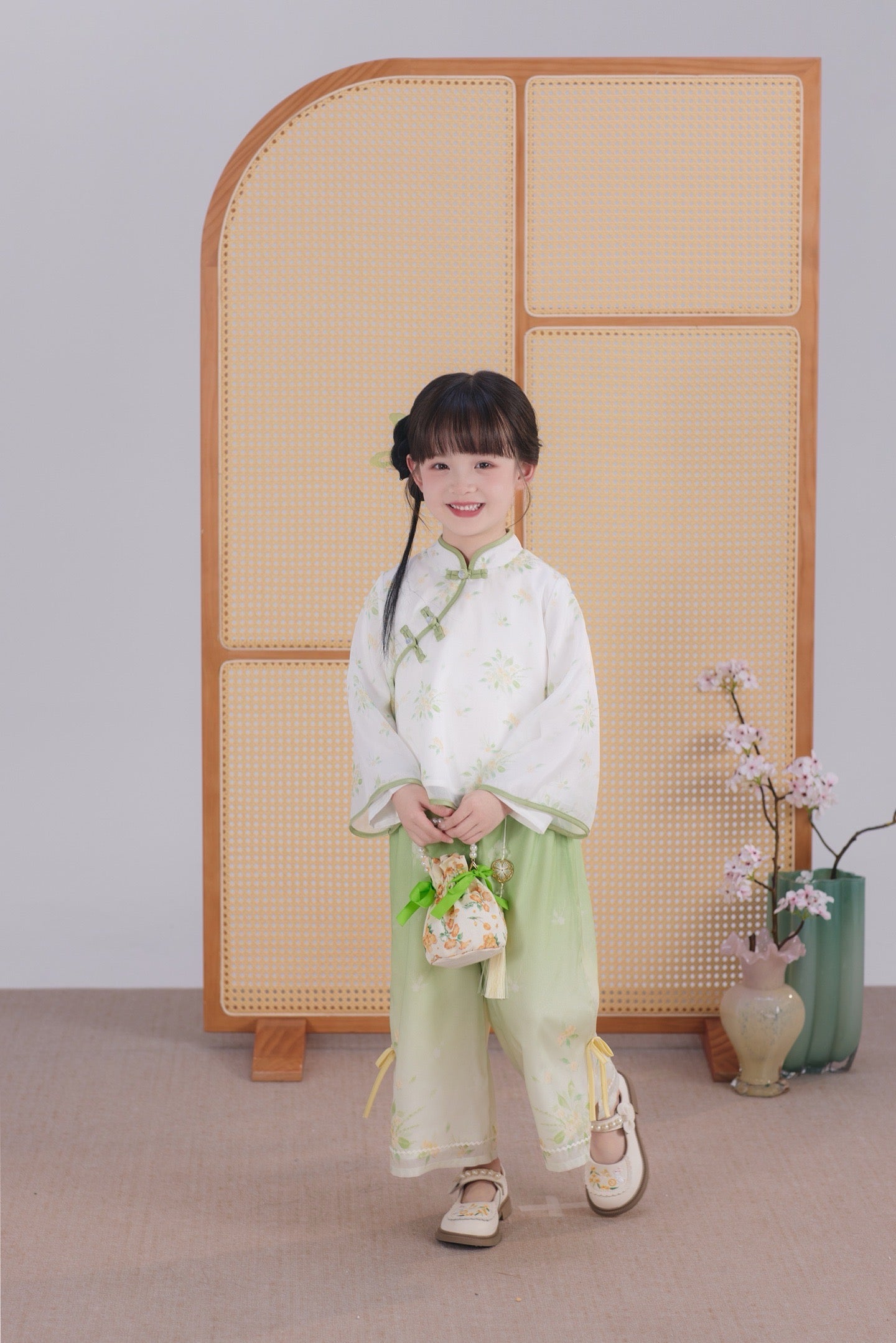 Child in traditional outfit standing in front of a woven screen with decorative vases.