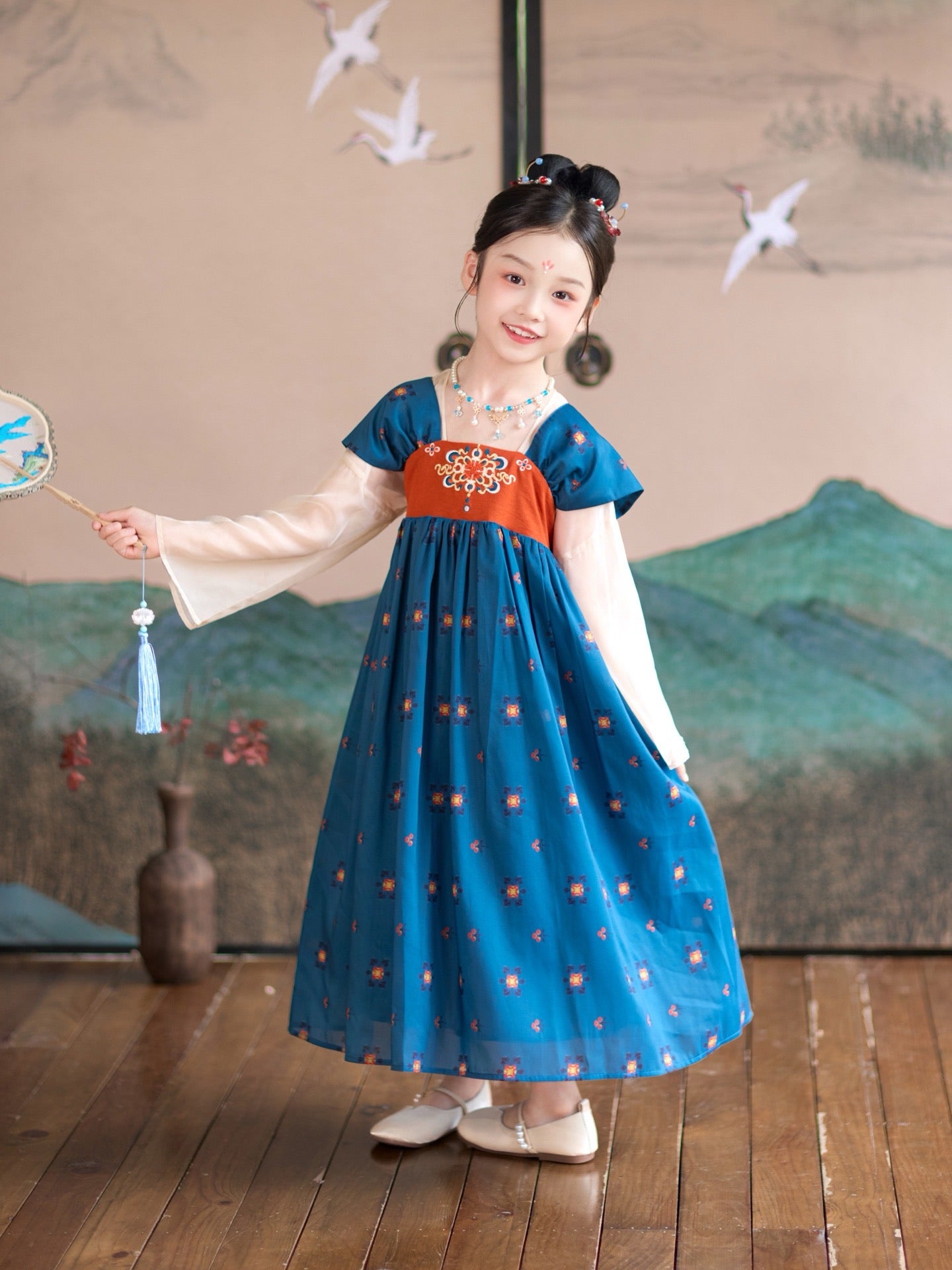 Young girl in a traditional outfit with a blue dress and red top, holding a fan, in a room with a scenic wall painting.