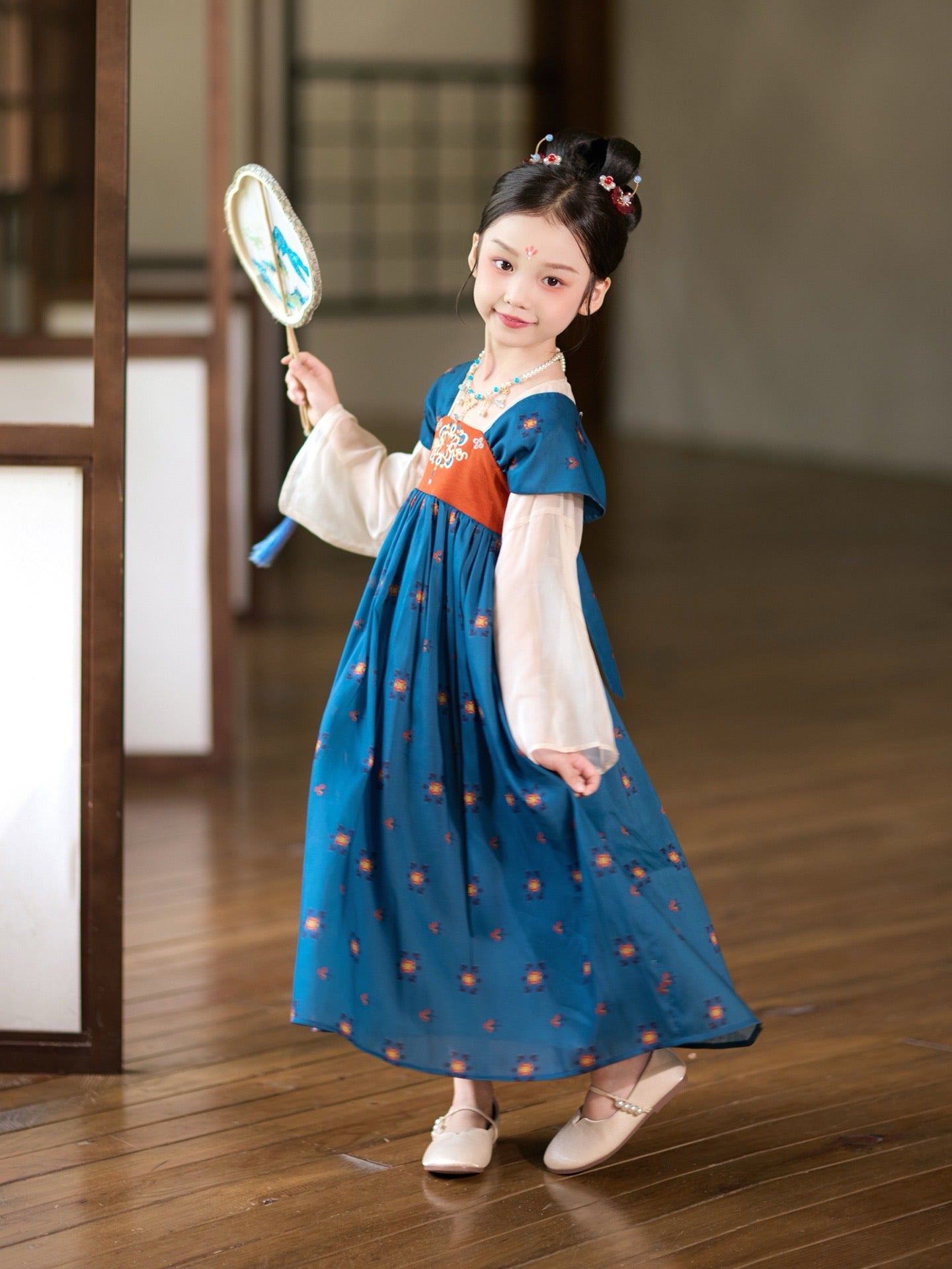 Young girl in a traditional outfit holding a fan on a wooden floor.