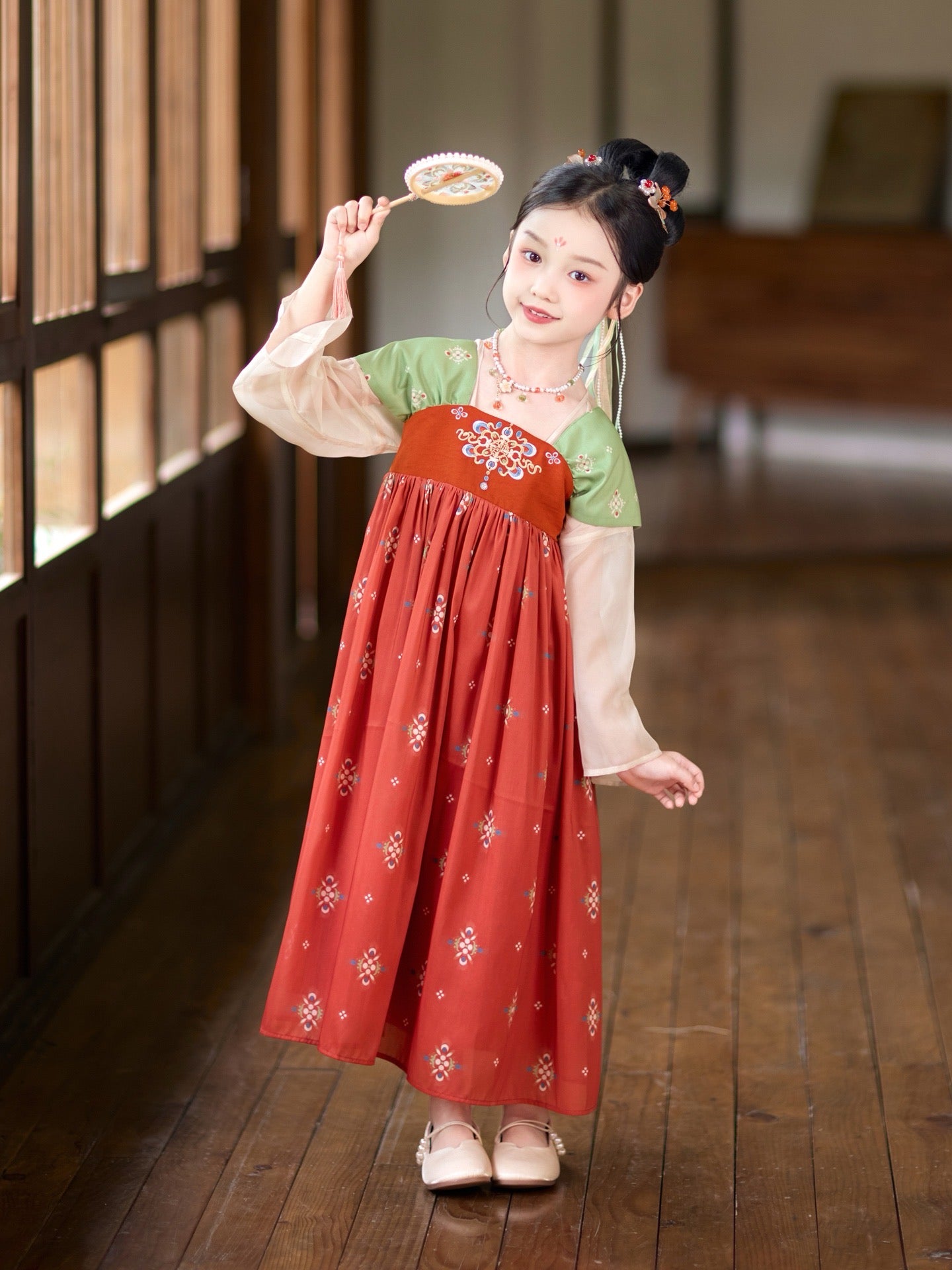 Young girl in a traditional outfit holding a fan in a room with wooden floors and bookshelves.