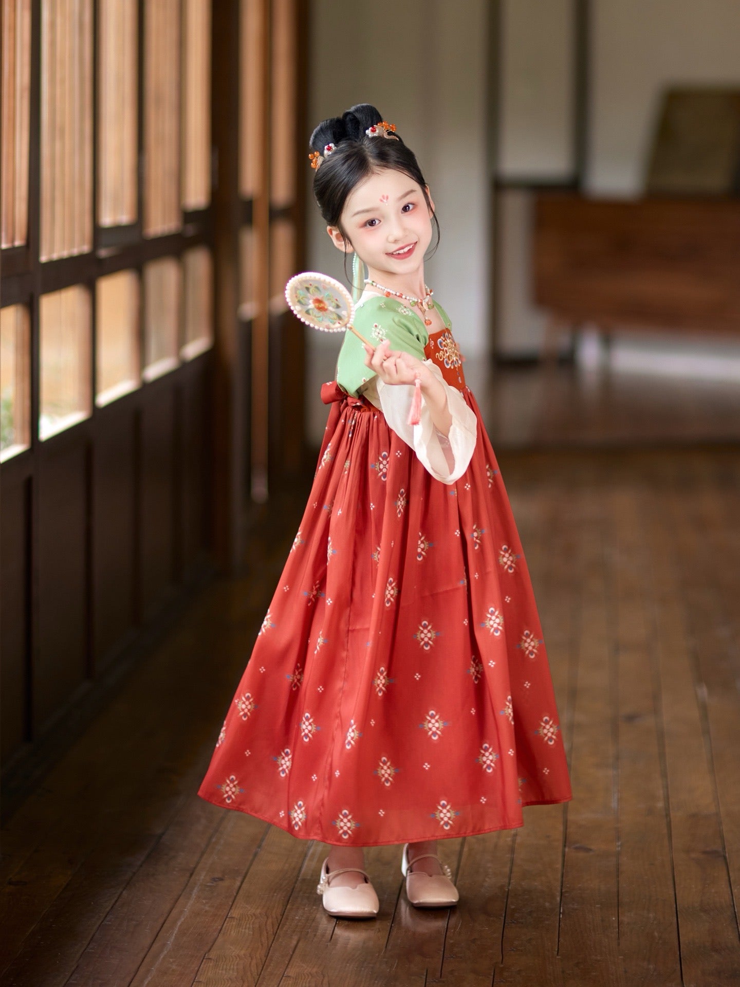 Child in traditional red and green dress holding a fan indoors.
